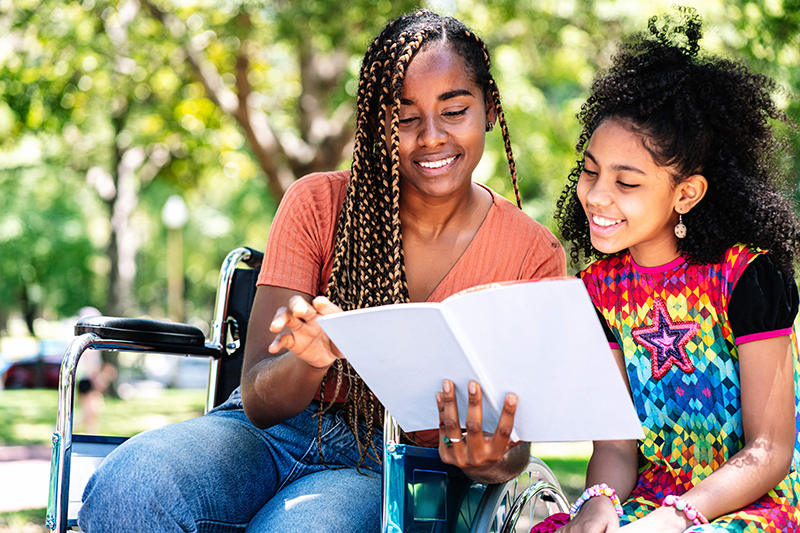 african-american-woman-wheelchair-enjoying-day-park-with-her-daughter-while-reading-book-together-min